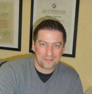 Ein Mann mit dunklem Haar und Bart trägt ein graues Oberteil und steht vor einer Wand mit einem gerahmten Zertifikat, das "Meisterbrief" enthält.