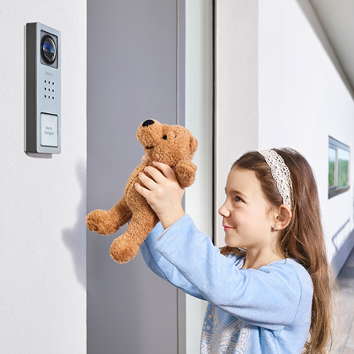 Ein kleines Mädchen hält einen Teddybären und steht vor einer Tür mit einem integrierten Video-Sprechanlage-System.